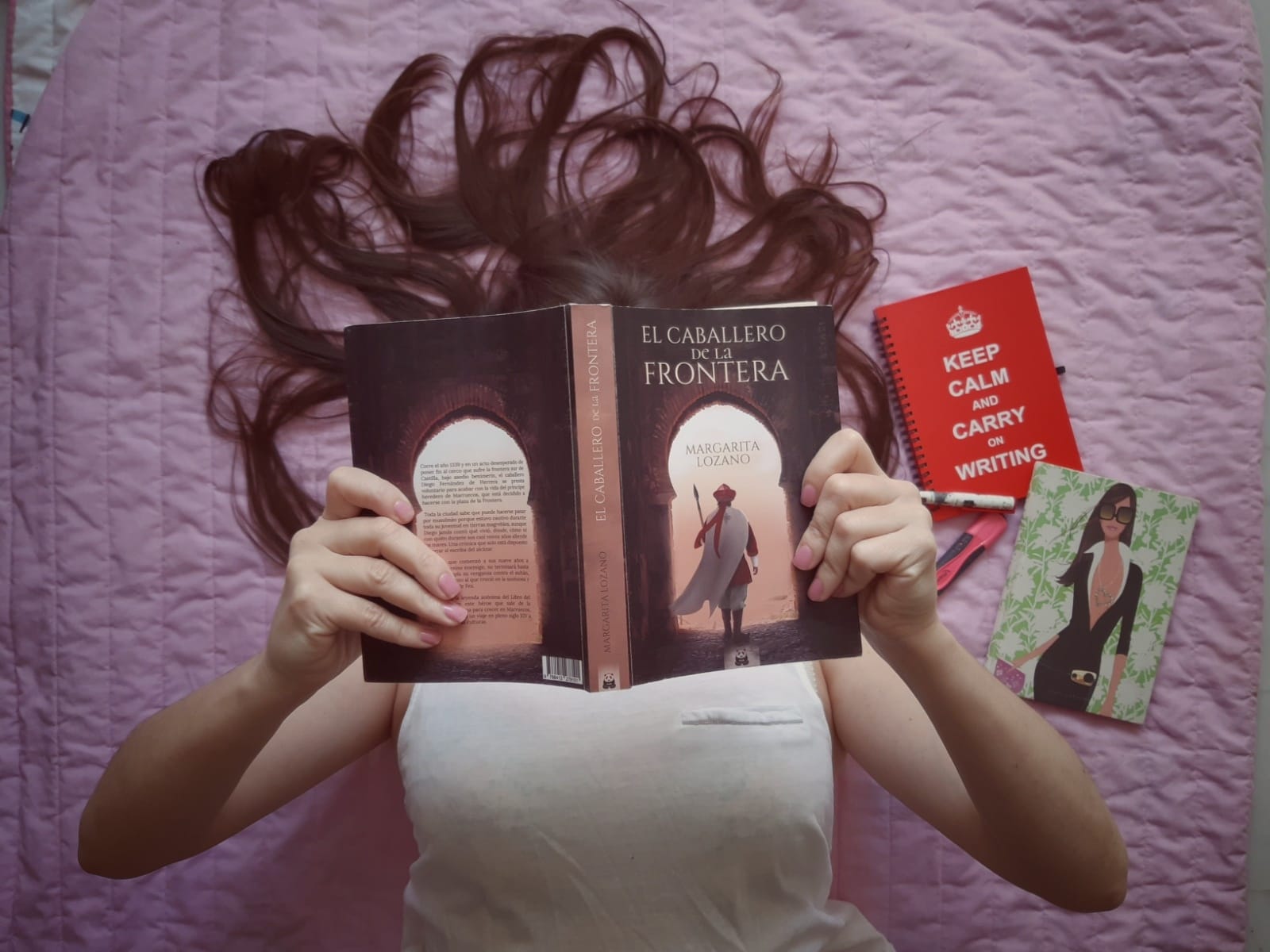 Mujer leyendo el libro "El caballero de la frontera" de Margarita Lozano, con cuadernos y bolígrafos sobre colcha rosa